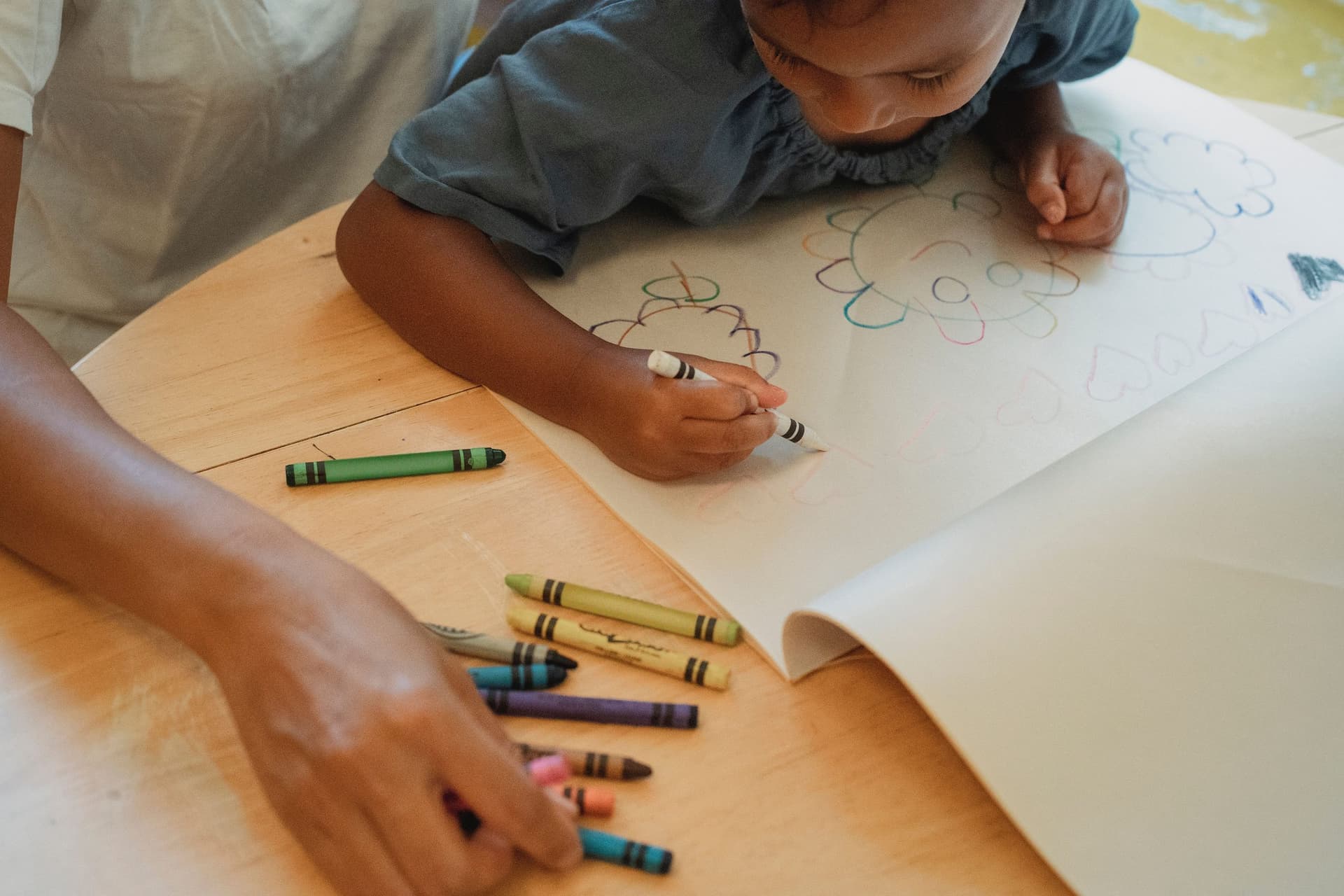 A young child draws with wax crayons on a large sheet of paper while an adult sorts the crayons