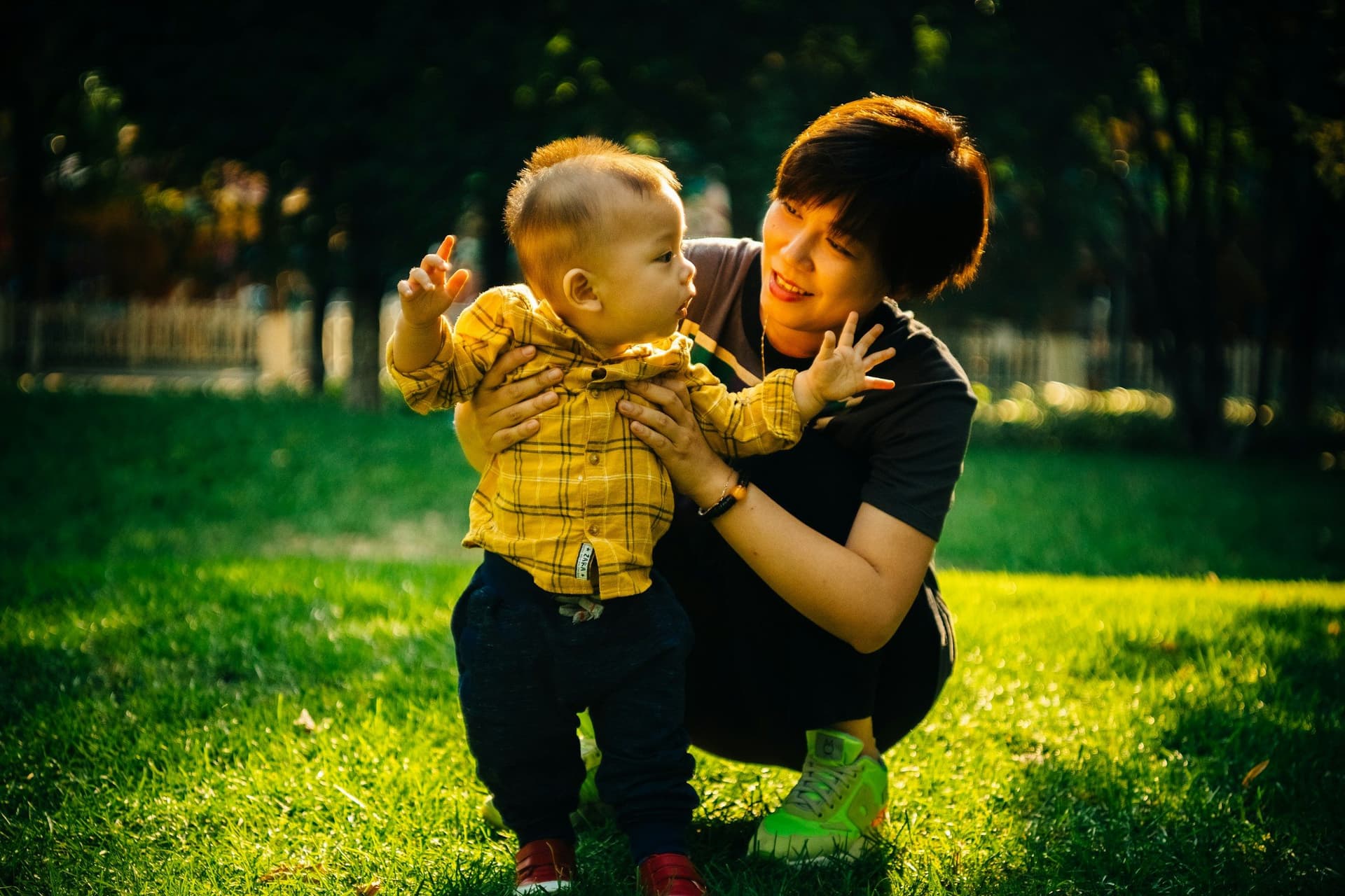 A woman crouches on sunlit grass in a park, steadying a toddler in a yellow checked shirt